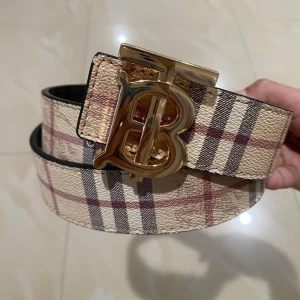 Burberry bälte - Snyggt bälte från Burberry med klassiskt rutigt mönster i beige, brun och röd. Bältet har ett stort guldigt B-spänne som verkligen sticker ut. Perfekt för att lyfta din outfit med en lyxig touch.