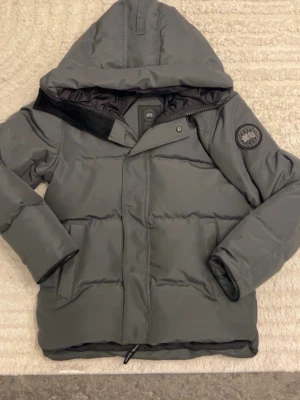 Grå pufferjacka från Canada Goose - Snygg grå pufferjacka från Canada Goose med huva och svart patch på ärmen. Jackan har dragkedja och tryckknappar framtill samt två sidofickor. Perfekt för kalla vinterdagar och riktigt varm tack vare det tjocka materialet.