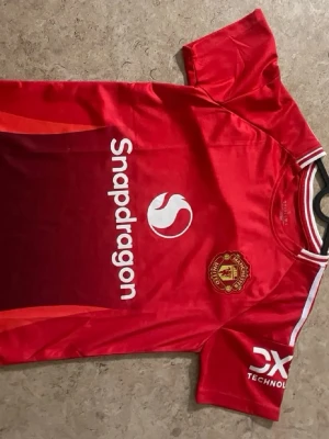 Manchester United röd matchtröja - Röd Manchester United matchtröja med Snapdragon-logga på bröstet och klubbemblem på vänster sida. Tröjan har vita detaljer vid halsen och på ärmen samt DXC Technology-logga på ena ärmen. Materialet är lätt och andas, perfekt för fotboll eller träning.
