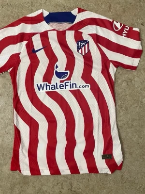 Atletico Madrid matchtröja Nike - Säljer en Atletico Madrid matchtröja från Nike med röda och vita vågiga ränder, klubbmärke på bröstet och sponsorloggor. Tröjan har korta ärmar och är tillverkad i lätt, ventilerande material som passar perfekt för fotboll och träning.