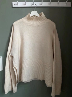 Beige oversized stickad polotröja - Mysig beige stickad polotröja med snygg passform och bred ribbstickning vid ärmslut och nederkant. Tröjan har hög krage och slitsar i sidorna för en avslappnad look. Perfekt för lager på lager under kyliga dagar.