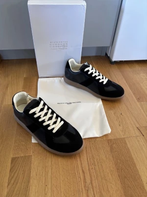 Maison Margiela svarta sneakers - Maison Margiela sneakers i svart läder med vita snören och beige gummisula. Klassisk low-top modell med stilren design och diskreta detaljer. Insidan är ljus och skon har en ikonisk sula med präglad logotyp. Perfekta för dig som gillar minimalistisk och exklusiv stil.