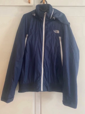 Blå vindjacka från The North Face - Snygg mörkblå vindjacka från The North Face med vit dragkedja och logga på bröstet och ryggen. Jackan har hög krage, två sidofickor med dragkedja och en luva. Materialet är lätt och vattenavvisande, perfekt för blåsiga dagar.