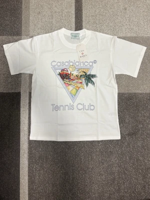 Casablanca Tennis Club t-shirt vit - Vit t-shirt från Casablanca med färgglatt tryck framtill som visar en tennisboll, palmer och geometriska mönster. Texten 'Casablanca Tennis Club' är tryckt på bröstet. Klassisk passform och rund hals. Perfekt för en sportig och trendig look.
