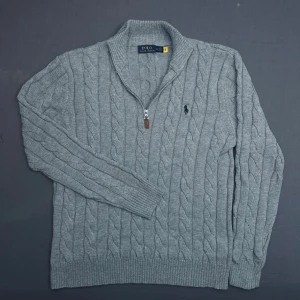 Grå kabelstickad halfzip Polo Ralph Lauren - Snygg grå kabelstickad tröja från Polo Ralph Lauren med halv dragkedja och hög krage. Tröjan har det klassiska broderade logotypen på bröstet och ribbade muddar. Perfekt för lager på lager och riktigt skön att bära. Skulle säga att tröjan passar som en stor M eller liten L