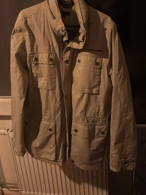 Beige vindjacka med fickor från dressman - Stilren beige vindjacka med fyra stora fickor framtill och knappdetaljer. Jackan har hög krage och dragkedja samt tryckknappar. Perfekt för dig som vill ha en enkel och funktionell jacka till vardags. Materialet känns lätt och smidigt. Storlek S men passar bäst på någon som är lite längre(M)