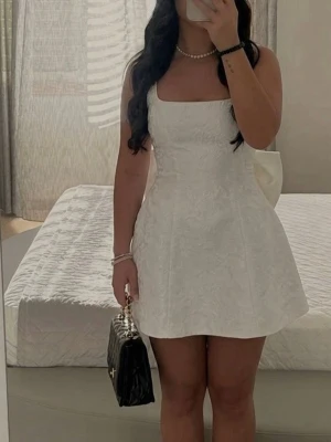 Vit kort klänning med stor rosett - Supersöt vit kort klänning med breda axelband och en stor rosett bak. Klänningen har en lätt utsvängd kjol och diskret struktur i tyget. Perfekt för dig som vill sticka ut med en unik detalj. Dragkedja bak för enkel påklädning.