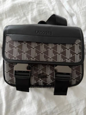 Lacoste väska  - Snygg handväska från Lacoste i brunt och svart med grafiskt mönster och vita detaljer. Väskan har ett lock med två spännen, justerbar axelrem och präglad Lacoste-logga på framsidan. Materialet är konstläder och textil, perfekt för dig som gillar streetstyle.