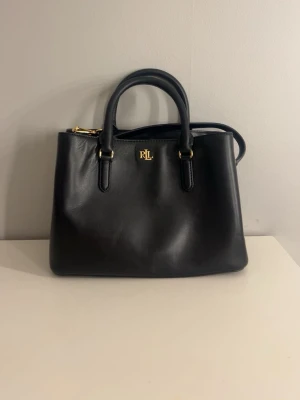 Svart handväska från Ralph Lauren - Stilren svart handväska från Ralph Lauren i slätt läder med guldfärgade detaljer och RL-logga framtill. Väskan har både handtag och en avtagbar axelrem för flexibel användning. Klassisk fyrkantig form som rymmer det viktigaste. Nyskick och aldrig använd. Nypris 4395 kr. Måtten är ungefär 29 cm x 13 cm och höjd 19 cm. Dustbag och kvitto finns.