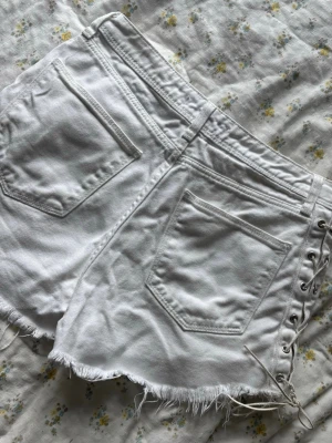 Vita shorts med snörning i sidan - Säljer ett par vita shorts med rå kant och snörning i sidan. De har fem fickor och en klassisk jeanslook med knapp och dragkedja framtill. 