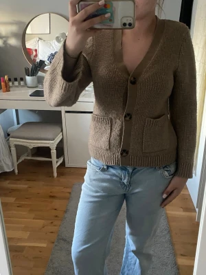 Beige stickad kofta med fickor - Mysig beige stickad kofta med v-ringning och stora knappar framtill. Koftan har två praktiska fickor och ribbade muddar vid ärmslut och nederkant. Perfekt att slänga över en t-shirt för en avslappnad look.