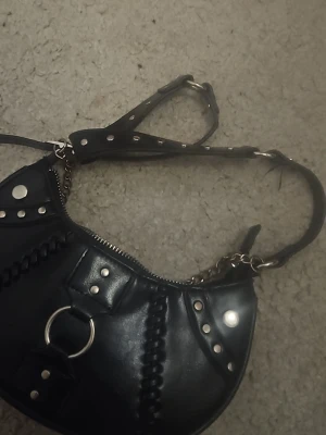 Svart axelväska med nitar och kedja - Cool svart axelväska i läderimitation med silverfärgade nitar och kedjedetaljer. Väskan har en rundad form, justerbar axelrem och dekorativa sömmar. Perfekt för dig som gillar edgy och trendiga accessoarer.