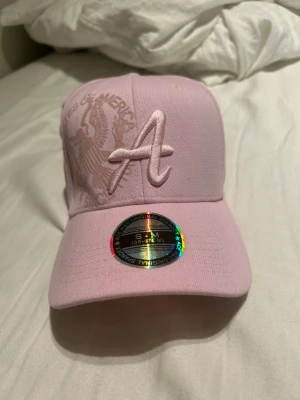 Ljusrosa keps från New Era - Säljer en ljusrosa keps från New Era med broderad 'A' framtill och diskret mönster på sidan. Kepsen har böjd skärm och klassisk passform. Tillverkad i mjukt bomullsmaterial och har justerbar baksida för perfekt fit.