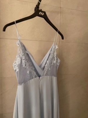 Ljusblå långklänning med spetsdetaljer - Elegant ljusblå långklänning med tunna axelband och v-ringning. Överdelen har fina spetsdetaljer och transparent mesh vid axlarna. Använd enbart en gång