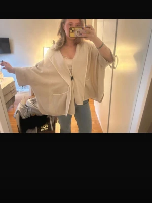 Beige poncho med knappar - Säljer en beige poncho med knapp framtill och två stora fickor. Ponchon har vida ärmar och en avslappnad passform, perfekt att slänga över en topp. Materialet ser mjukt och bekvämt ut, och färgen är lätt att matcha med det mesta.