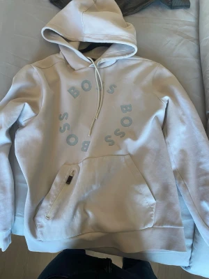 Vit hoodie från BOSS med tryck - Snygg vit hoodie från BOSS med cirkulärt tryck på bröstet. Hoodien har en stor ficka med dragkedja och snörning i huvan. Perfekt för dig som gillar stilrena och moderna plagg.