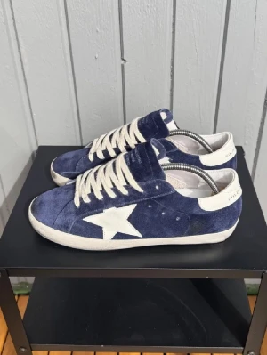 Blåa Golden Goose Superstar sneakers - Snygga blåa Golden Goose Superstar sneakers med vit stjärna på sidan och vita detaljer som snörning och häl. Skorna är gjorda i mocka och har en klassisk låg profil som passar perfekt till jeans och streetwear.
