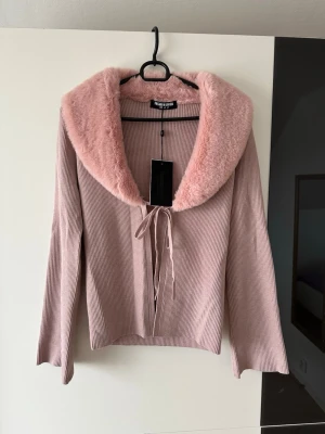 Rosa kofta med fuskpäls Fashion Union - Supersöt ljusrosa ribbad kofta från Fashion Union med stor krage i mjuk fuskpäls. Koftan har långa ärmar och knyts med ett band framtill. Perfekt för dig som vill ha en trendig och mysig look.