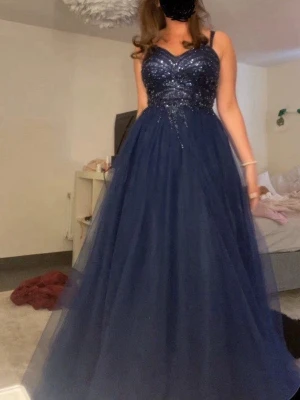 Marinblå glittrig bal klänning  - Elegant marinblå långklänning med glittrig överdel och tunna axelband. Kjolen är i flera lager tyll som ger en svepande känsla. Perfekt för bal eller festliga tillfällen. Snygg hjärtformad urringning och markerad midja.
