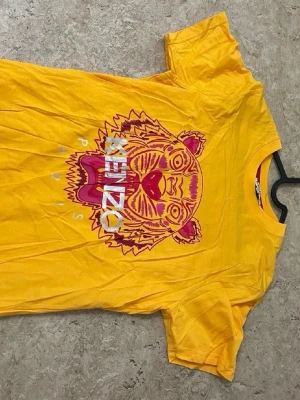 Gul Kenzo t-shirt med tigertryck - Cool gul t-shirt från Kenzo med ett stort tigertryck i rosa och lila på bröstet. T-shirten har rund hals och korta ärmar, perfekt för dig som vill sticka ut. Loggan är tryckt över motivet och materialet känns mjukt och bekvämt.