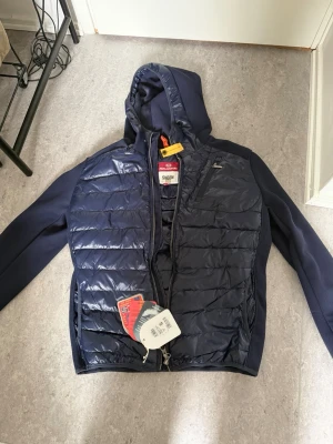 Parajumper cardigan - Mörkblå cardigan från parajumper med huva och quiltad front. Jackan har dragkedja och är tillverkad i ett glansigt material med paneler av stretchtyg på ärmar och sidor. Perfekt nu inför våren och snygg att matcha med jeans.