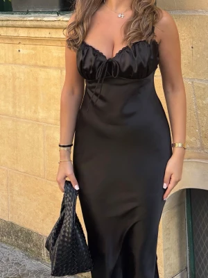 Svart långklänning med snörning - Elegant svart långklänning i satin med smala axelband och snygg snörningsdetalj vid bysten. Klänningen har en figurnära passform och en mjukt skimrande yta som ger en lyxig känsla. Perfekt för dig som vill ha en stilren och trendig look.
