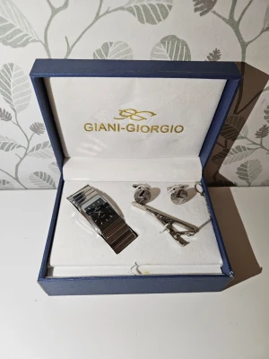 Giani-Giorgio silverfärgad klockset vintage  - Snyggt set från Giani-Giorgio med en stilren silverfärgad klocka med svart urtavla, matchande manschettknappar och slipsnål. Klockan har en bred länk i metall och en rektangulär form på urtavlan. Perfekt för dig som gillar klassisk och elegant stil. Perfekt för egen användning eller present. Klocka manscettknappar och slipsnål är i nyskick och låda är i bra skick