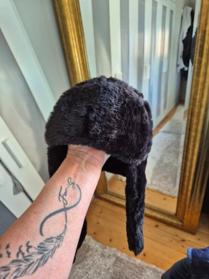Svart pälsmössa med öronlappar - Mysig svart pälsmössa med öronlappar och spänne under hakan. Hatten är gjord i mjuk fuskpäls och har en klassisk trappermodell som håller dig varm under kalla dagar. Perfekt accessoar för vintern och riktigt snygg till streetwear.