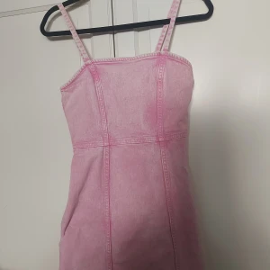 Rosa jeansklänning med smala axelband - Supersöt rosa kort jeansklänning med smala axelband och markerade sömmar. Klänningen har en dragkedja i ryggen som gör den enkel att ta på. Perfekt för dig som gillar en cool och trendig look med denimkänsla.