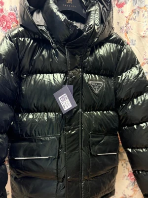 Svart glansig dunjacka från Prada - Säljer en svart, glansig dunjacka från Prada med huva och stora framfickor. Jackan har en klassisk puffer-modell, dragkedja och tryckknappar framtill samt Pradas ikoniska trekantslogga på bröstet. Perfekt för kalla vinterdagar och riktigt snygg streetstil.