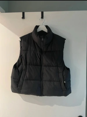 Svart dunväst från 157 - Snygg svart dunväst från 157 med hög krage och dragkedja framtill. Västen har två sidofickor och quiltad design som ger en schysst look. Perfekt att slänga över en hoodie eller tröja när det är kyligt ute. Materialet är mjukt och puffigt för extra värme.
