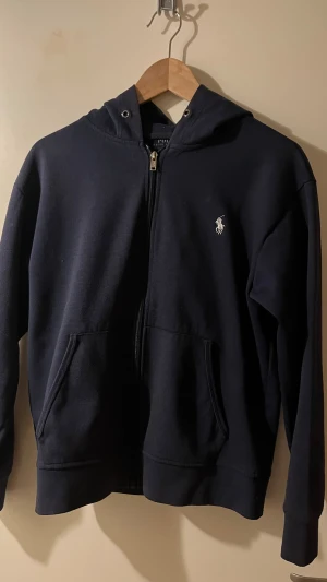 Marinblå hoodie från Polo Ralph Lauren - Hej!       Säljer en snygg hoodie från Polo Ralph Lauren med dragkedja och huva.           Tröjan har klassisk logga broderad på bröstet och praktiska fickor framtill.             Tillverkad i mjuk bomull och polyester för skön komfort.  Perfekt för chill dagar eller när du vill ha en clean look.                                                           Vill du veta något mer eller vill ha fler bilder så är det bara att skriva! Priset är såklart inte hugget i sten!