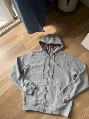 Grå hoodie från Burberry - Säljer en grå zip-hoodie från Burberry med klassiskt rutigt foder i huvan och broderad logga på bröstet. Hoodien har dragsko i huvan, två fickor fram och ribbade muddar. Perfekt för en chill och stilren look. Storleken är XL men det kan kännas som ett L eller M 