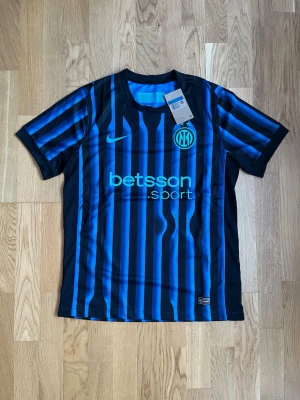 Randig Inter fotbollströja Nike - Säljer en svart och blå randig Inter fotbollströja från Nike med klubbmärke och sponsortryck på bröstet. Tröjan är kortärmad och tillverkad i lätt polyester, perfekt för fotboll eller träning. Klassisk sportig design med moderna detaljer.