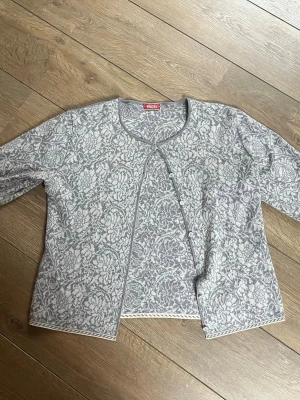 Gråmönstrad kofta från Oleana - Grå kofta med ljusgrått blommönster från Oleana. Koftan har rund hals, knäppning framtill med små knappar och långa ärmar. Mjuk och skön ullblandning med fina detaljer vid muddar och nederkant. Har en jätteliten gul fläck på framsidan som visas på bild 3. Fråga Järna vid frågor 💕