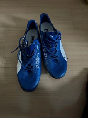 Blåa Puma Ultra fotbollsskor - Säljer ett par blåa Puma Ultra fotbollsskor med vita detaljer och texten 'ULTRA' och 'SPEED' på ovansidan. Skorna har snörning, låg profil och dobbar för bra grepp på planen. Tillverkade i syntetmaterial för lätt känsla och snabbhet.