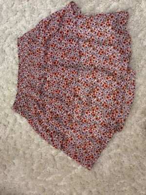 Blommig kortkjol i rött och vitt - Supersöt kortkjol med blommigt mönster i rött, vitt och lite grönt. Kjolen har en avslappnad passform och resår i midjan för extra komfort. Perfekt för vår och sommar, lätt att matcha med en stickad tröja eller t-shirt.