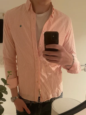 Randig skjorta från Morris Stockholm - Säljer en snygg, vit och ljusorange randig skjorta från Morris Stockholm i storlek M. Skjortan har klassisk krage, långa ärmar och knappar framtill. Tillverkad i mjuk bomull och har en liten grön broderad detalj på bröstet.