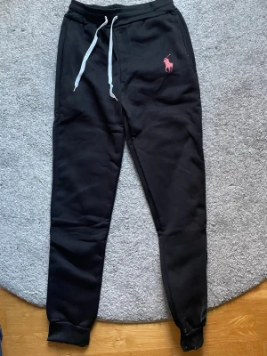 Ralph Lauren mjukisbyxor - Svarta mjukisbyxor från Ralph Lauren med vit dragsko i midjan och rosa broderad logga på vänster sida. Var du frågor så bara hör av dig!