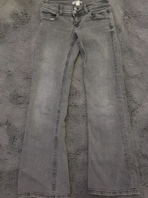Grå bootcut jeans med stjärndetalj - Säljer ett par grå bootcut jeans med klassisk femficksmodell och en snygg stjärndetalj på ena bakfickan. Jeansen har en lätt tvättad look och raka, något utsvängda ben. Perfekta för dig som gillar en avslappnad men trendig stil.