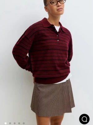 Vinröd randig polotopp med knappar - Vinröd polotopp med svarta ränder och knappar framtill. Klassisk krage och lång ärm, perfekt för en avslappnad men ändå stilren look. Mjuk och bekväm, passar till både kjol och byxor.