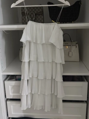 Vit volangklänning med tunna band - Säljer en superfin vit klänning med tunna axelband och flera lager volanger. Klänningen är kort och har en luftig, plisserad design som ger en romantisk och trendig känsla. Perfekt för sommarens alla tillfällen!