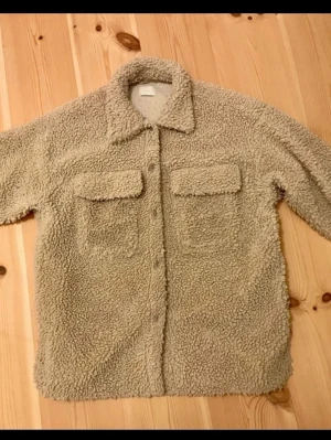 Beige teddyjacka med fickor - Mysig beige teddyjacka i fluffigt material med två stora bröstfickor och knappar framtill. Jackan har klassisk krage och avslappnad passform, perfekt för lager på lager. Passar dig som gillar en chill och trendig stil.