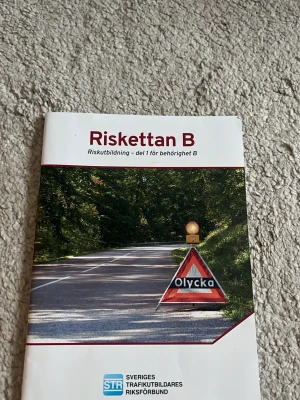Riskettan B kursbok STR - Kursbok för Riskettan B, del 1 för behörighet B. Boken tar upp riskutbildning för bilförare med fokus på alkohol, droger, trötthet och riskfyllda beteenden i trafiken. Omslaget är vitt med en vägskylt och text. Perfekt för dig som pluggar till körkort.
