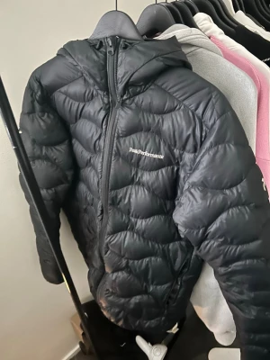 Svart dunjacka Peak Performance - Snygg svart dunjacka från Peak Performance med huva och dragkedja framtill. Jackan har diskret logga på bröstet och quiltad design som ger en sportig look. Perfekt för kalla vinterdagar tack vare det värmande dunmaterialet.