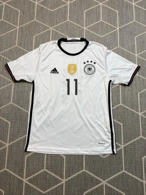Tysklands Adidas  - Vit matchtröja från Adidas för Tysklands landslag med nummer 11, svart krage och detaljer, samt FIFA-märke och fyra stjärnor på bröstet. Tillverkad i lätt och ventilerande material, perfekt för fotbollsträning eller match.
