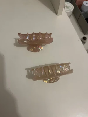Två pärlemorskimrande hårklämmor - Säljer två snygga hårklämmor i pärlemorskimrande ljusrosa plast. De har en klassisk rektangulär form med stadiga grepp och passar perfekt för att hålla upp håret på ett trendigt sätt. Perfekt accessoar för att piffa till frisyren snabbt.