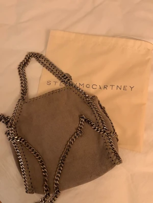 Grå Stella McCartney Falabella väska tiny - Snygg grå handväska från Stella McCartney i modellen Falabella. Väskan har kedjedetaljer längs kanterna och dubbla kedjehandtag. Tillverkad i veganskt läder med en mjuk, lyxig känsla och cool design. Perfekt för dig som gillar statement-accessoarer. Väskan har defekt men inger jag tycker märkt när man bär den. Priset kan diskuteras vid snabb affär!!