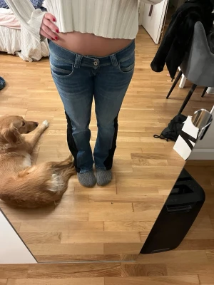 Blå jeans med svarta detaljer - Säljer ett par blå jeans med svarta paneler längs benen. Jeansen har låg midja, klassisk femficksmodell och raka ben. Materialet är denim med stretch för extra komfort. Perfekta för en trendig och avslappnad look.