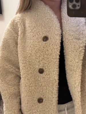 Beige teddyjacka från Gina Tricot - Supermysig beige teddyjacka från Gina Tricot med stora bruna knappar framtill. Jackan har en fluffig och mjuk struktur som ger en riktigt cozy vibe. 